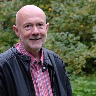 Markus Kaminski schaut in die Kamera, im Hintergrund sind grüne Büsche zu sehen. Er trägt ein rot-weiß kariertes Hemd mit einer dunkeln