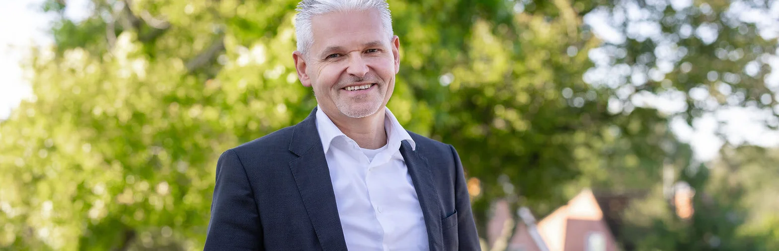 Ein Mann mit kurzen grauen Haaren lächelt in die Kamera. Er trägt ein helles Hemd und ein dunkles Sakko.